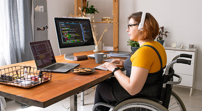 Female employee working on laptop and large screen monitor, wearing headphones, sitting in wheelchair.